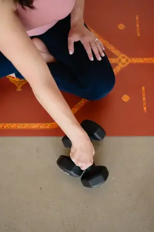Tracy H. on yoga mat picking up dumbbells during fitness session.